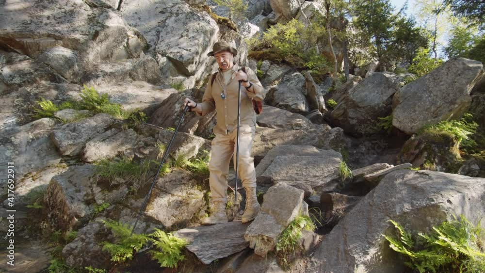 Low angle view of senior male tourist with backpack walking down rocky mountain trail with trekking poles while hiking in wild nature on summer day Low angle view of senior male tourist with backpack walking down rocky mountain trail with trekking poles while hiking in wild nature on summer day