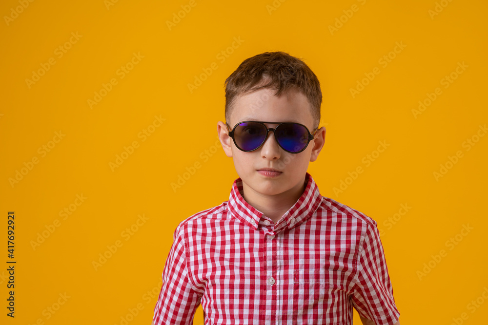 stylish boy in dark sunglasses and a red checked shirt looks at the camera, standing against the yellow background of the studio. Observes the environment