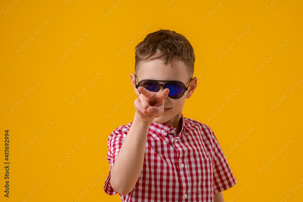 suspicious boy in dark sunglasses and a red checked shirt looks doubtfully at the camera., standing against the yellow background of the studio. Observes the environment suspicious boy in dark sunglasses and a red checked shirt looks doubtfully at the camera., standing against the yellow background of the studio. Observes the environment