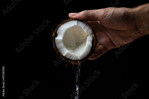 Isolated coconut on a black background. Coconut milk is poured from half a coconut. Half of a coconut in a man's hand on a dark background. Healthy fruit