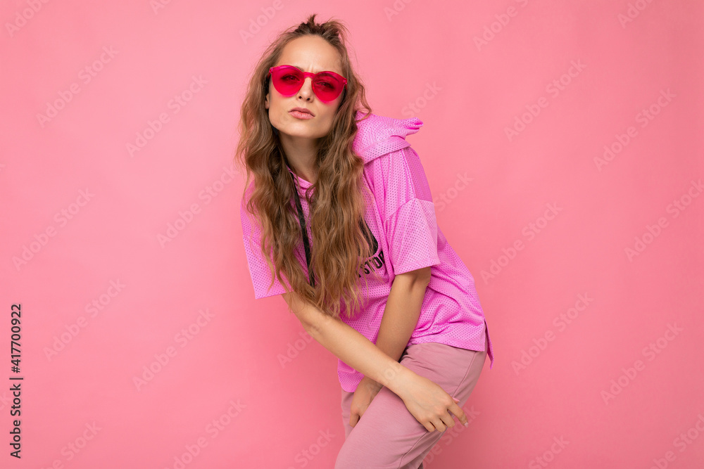 Photo shot of beautiful young dark blonde woman wearing casual clothes and stylish sunglasses isolated over colorful background looking at camera Photo shot of beautiful young dark blonde woman wearing casual clothes and stylish sunglasses isolated over colorful background looking at camera