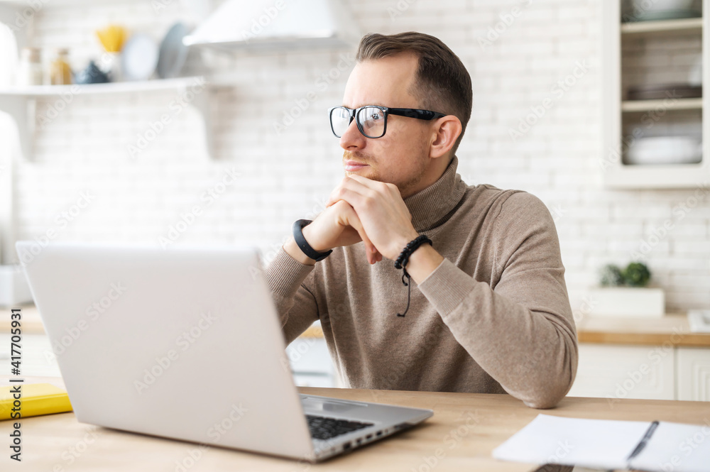 Confident male worker on the break, at the laptop, looking away, holding hands near the chin, thinking about plans, vacation, weekends, remembered a nice memory, home office, dressed in beige sweater Confident male worker on the break, at the laptop, looking away, holding hands near the chin, thinking about plans, vacation, weekends, remembered a nice memory, home office, dressed in beige sweater