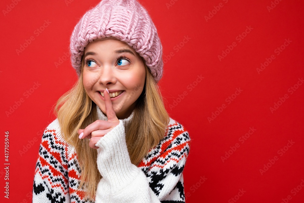 Closeup of young positive happy beautiful blonde woman with sincere emotions wearing pink hat and winter sweater isolated over red background with copy space and showing silence gesture with finger Closeup of young positive happy beautiful blonde woman with sincere emotions wearing pink hat and winter sweater isolated over red background with copy space and showing silence gesture with finger