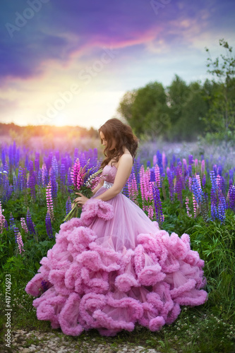 A young girl in a lush pink ball gown with a wild flowers bouquet in her hands against the background of a blooming purple and pink lupines field and a sunset sky.