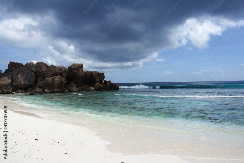 Seychelles, La Digue, Anse Cocos Beach, red granite rocks in the back with white sandy beach and turquoise blue sea in front, blue sky with big dark rain cloud