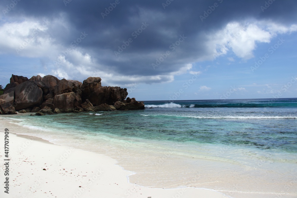 Seychelles, La Digue, Anse Cocos Beach, red granite rocks in the back with white sandy beach and turquoise blue sea in front, blue sky with big dark rain cloud Seychelles, La Digue, Anse Cocos Beach, red granite rocks in the back with white sandy beach and turquoise blue sea in front, blue sky with big dark rain cloud