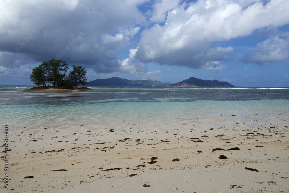 Seychelles, La Digue, Anse Reunion, white sandy beach with shallow turquoise blue water of coral reef small island with trees, white waves and bigger island in the back, blu sky and white clouds Seychelles, La Digue, Anse Reunion, white sandy beach with shallow turquoise blue water of coral reef small island with trees, white waves and bigger island in the back, blu sky and white clouds