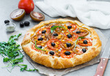 Galette with red and black tomatoes, olives, arugula and ricotta on paper on a light background 