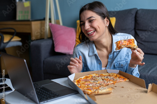 A young woman is eating pizza and having an online call on her laptop.