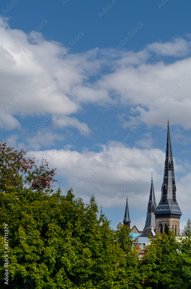 Trio of historical church steeples pointing to the cloudy blue sky above the treeline.