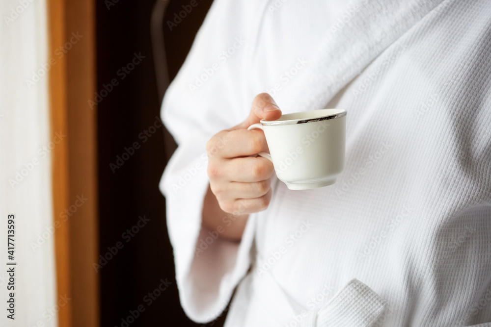 A man in a white terry dressing gown holds a coffee mug in front of a window. A man in a white terry dressing gown holds a coffee mug in front of a window.