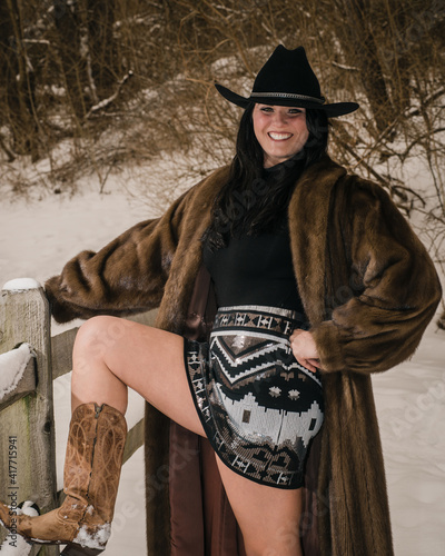 woman in fur coat and cowboy hat leaning against snow covered fence