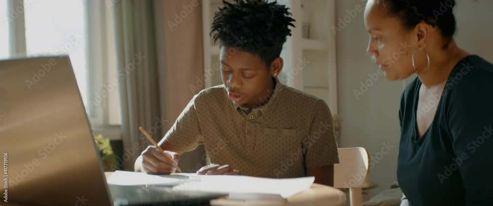 African American Black mother sits near her son, helping him with homework. Distance learning from home during COVID-19 coronavirus pandemic, stay home concept. Shot with 2x anamorphic lens