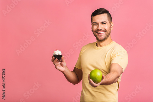 Joyful young Caucasianman in casual made wrong choice deciding between cake and fresh green apple. People, health, food, dieting and calories concept isolated over pink background. photo