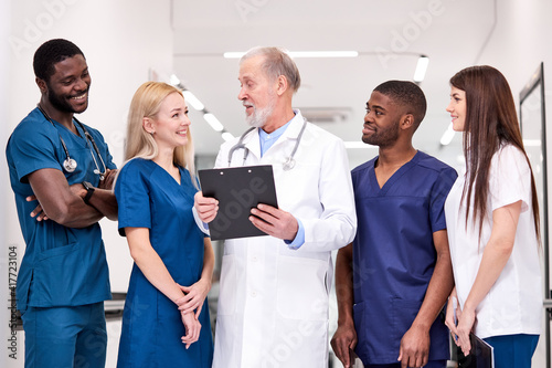 Positive senior doctor in white uniform and young doctors therapists posing looking at camera, elderly man holding clipboard in hospital, multidisciplinary medical team have talk