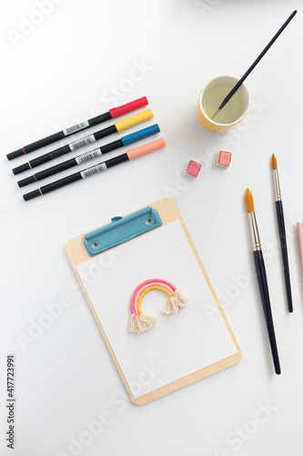 Stationery utensils over white background. A clipboard with a rainbow at the top are in the middle.