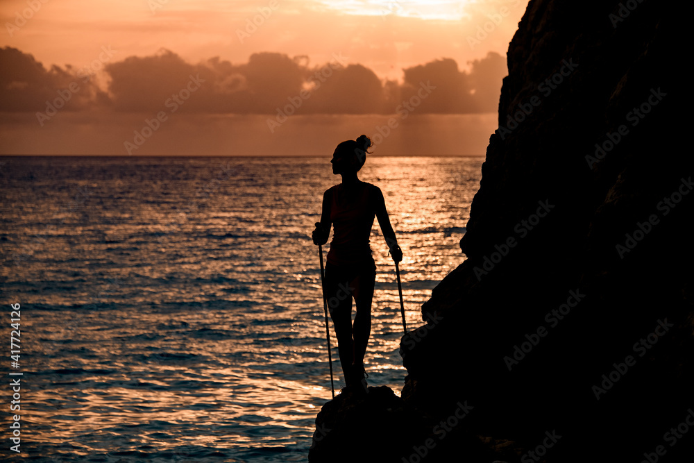 amazing view of female silhouette with trekking sticks near cliff on background with sunrise