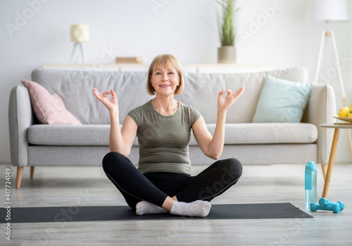 Balanced senior woman sitting in lotus pose and making gyan mudra with her hands, meditating on sports mat at home