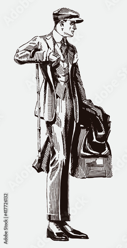 Standing stylish man from the early 20th century wearing suit, cap, gloves and gaiters, holding travel bag, overcoat and walking stick