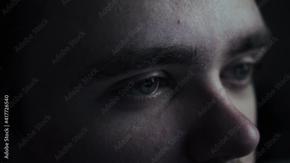 A close-up of a young guy sitting in a dark room watching TV with fright and fear. Focus on one side of the face. Weekend and relaxation concept A close-up of a young guy sitting in a dark room watching TV with fright and fear. Focus on one side of the face. Weekend and relaxation concept