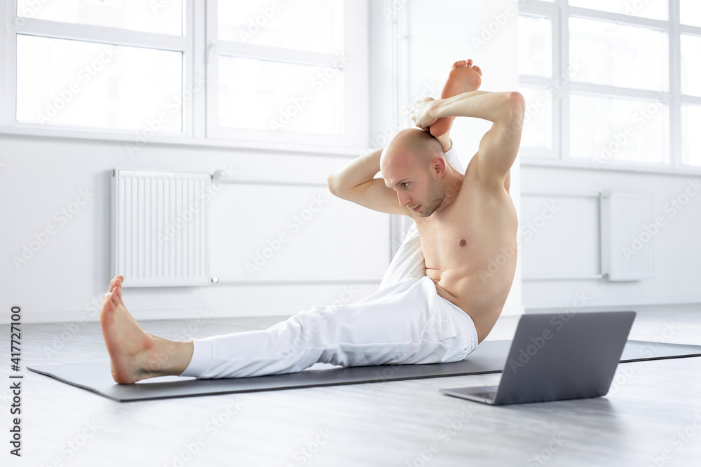 Naklejka premium shirtless yogi man showing yoga exercises at laptop camera, teaching online via Net, webcam. man sitting on floor , flexible and sportive