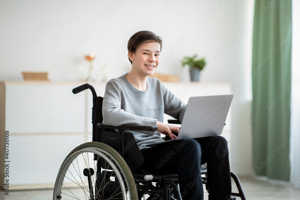 Happy disabled teenager in wheelchair using laptop computer for online studies at home Happy disabled teenager in wheelchair using laptop computer for online studies at home