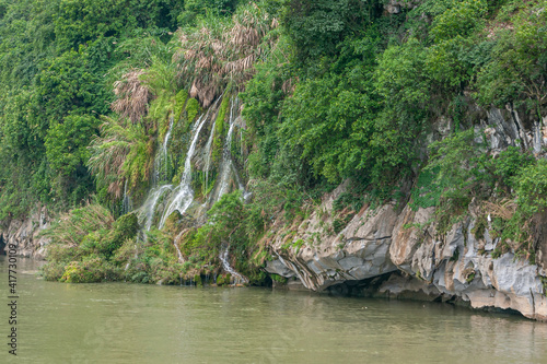 Guilin, China - May 10, 2010: Along Li River. Landscape, White Waterfalls over cave at green water level in beige rock cliff partly covered by green trees. 