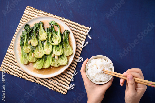 Stir fried bok choy with soy sauce on plate eating with cooked rice by using chopsticks on blue background, Asian vegan food