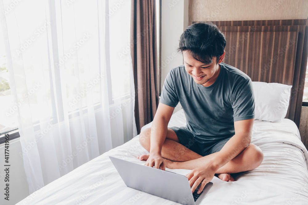 Happy Asian man is working with his laptop on his bed. Concept of freelancer successful lifestyle.