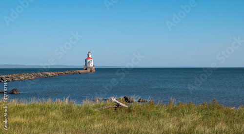 Lighthouse with red roof on Lake Superior at the end of a pier, large boulders in blue water, green grass and driftwwood, on a sunny afternoon.