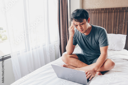 Tired stressed young Asian man feeling sleepy and tired while using laptop on the bed in bedroom. Hard work concept. © Reezky