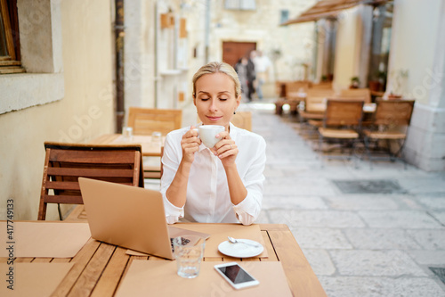 Technology and travel. Working outdoors. Freelance concept. Pretty young woman using laptop and drinking cofee in sidewalk cafe on ancient europian street.