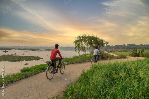 Fotografija Men riding bicycles on unpaved village road beside a river at West Bengal, India