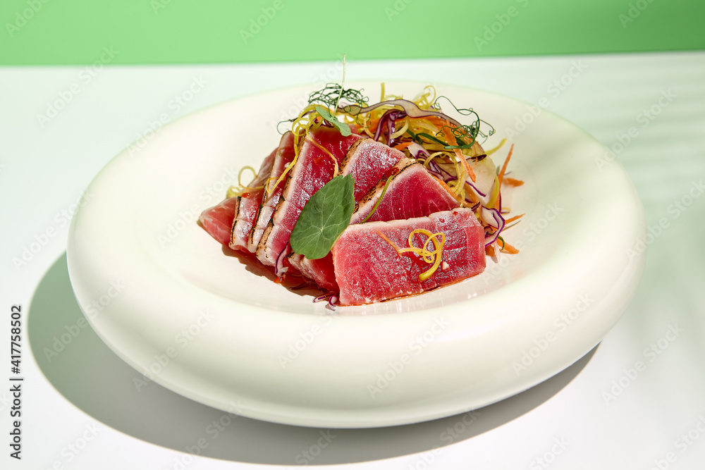 Tuna tataki in sesame with salad. Fish sashimi restaurant appetizer on white table with green wall. Day sunlight with hard shadow of tropical palm leaves. Summer or spring food concept. Tuna tataki in sesame with salad. Fish sashimi restaurant appetizer on white table with green wall. Day sunlight with hard shadow of tropical palm leaves. Summer or spring food concept.