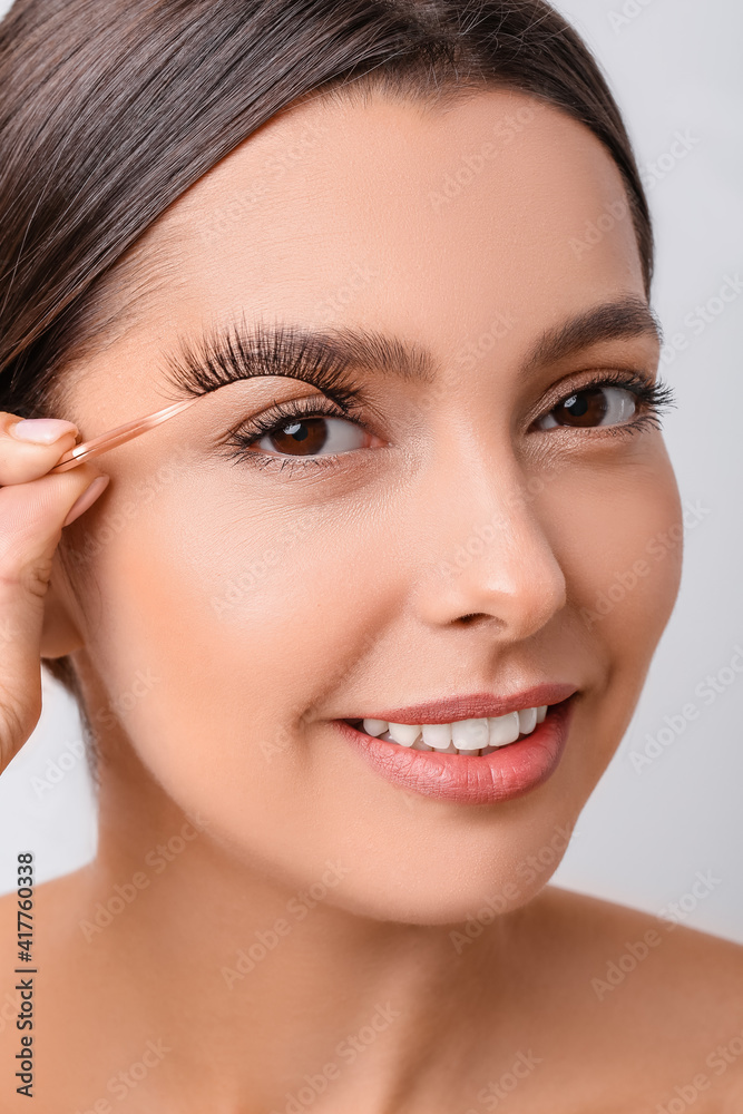 Beautiful young woman applying fake eyelashes against light background