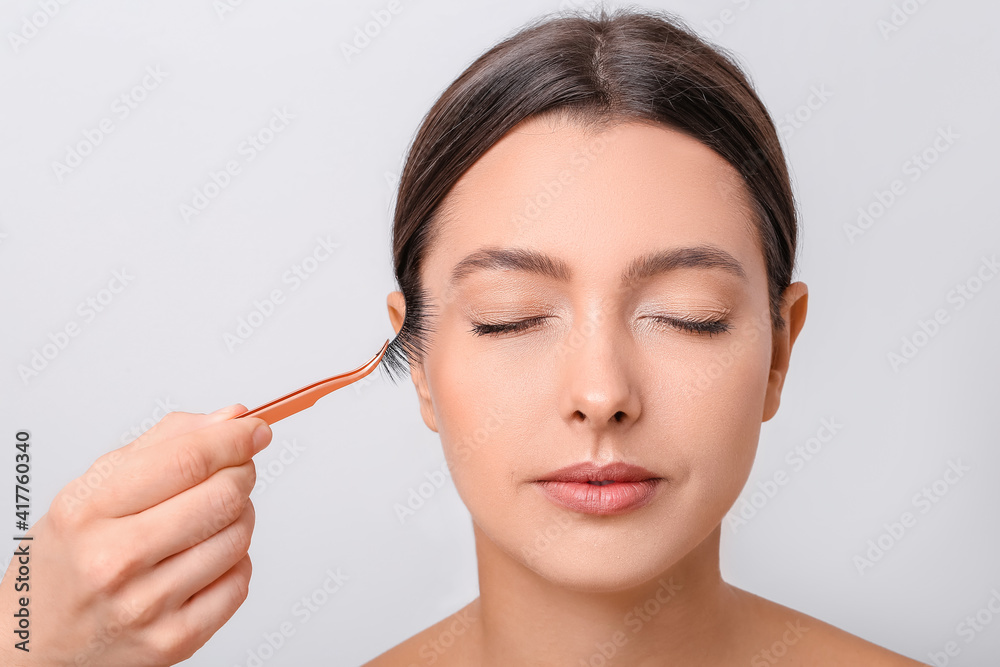 Beautiful young woman applying fake eyelashes against light background Beautiful young woman applying fake eyelashes against light background