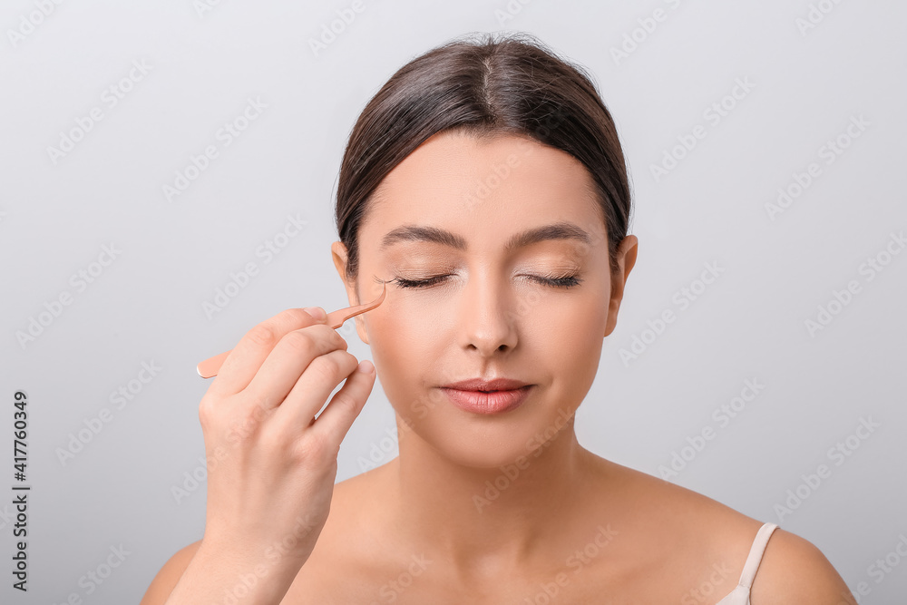 Beautiful young woman applying fake eyelashes against light background