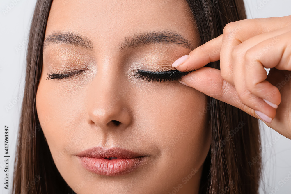 Beautiful young woman applying fake eyelashes against light background