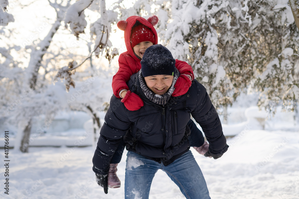 Young Caucasian dad is rolling his 6 year old daughter on his back in a winter park. Snow and family games in winter.