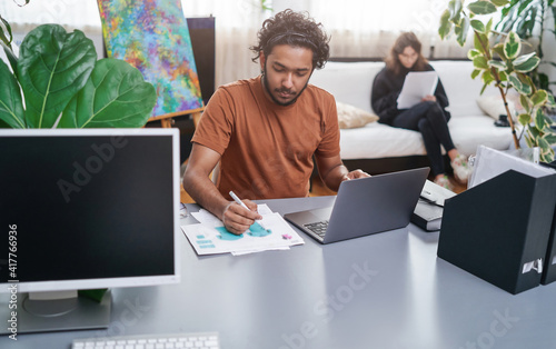 Cheerful male designer with curly hairs in casual attire sits at table and designing concept of shirts using a laptop and a pen.