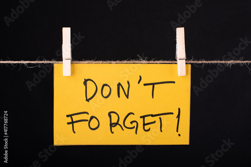 Don't Forget word written on a Yellow color sticky note hanging with a wire on black background.