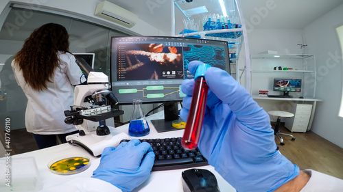 POV of virologist doctor holding tube with blood sampe typing information in pc, analysing patient disease, examining virus evolution. Team of scientists doing research working in equipped laboratory. photo