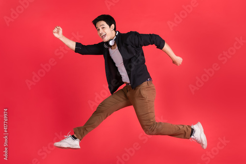 Asian man high jumping in cheerful and excited gesture with headphone on his neck. Studio light shot on red background In the concept of listening to fun music. © Bangkok Click Studio