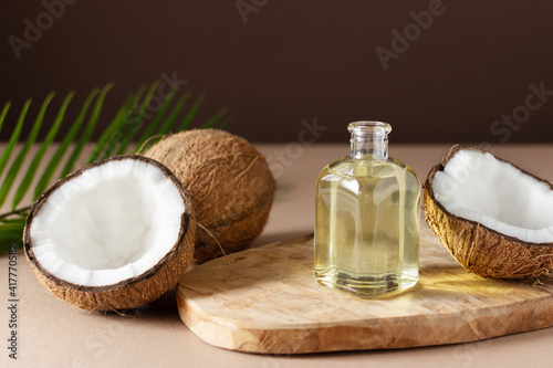 Bottle of coconut oil and fresh coconuts with palm leaf on wooden board over brown background.