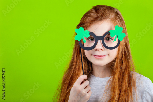 Portrait of little smiling caucasian red-haired girl in mask of leprechaun shamrock clover glasses for irish St. Patrick's Day on green studio background. Copyspace.