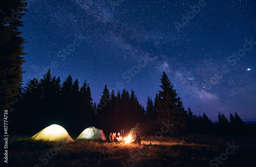 Company of five hikers, men and woman sitting by burning bonfire near tourist tents, enjoying beautiful camping night under dark sky full of stars and bright Milky Way, warm summer evening.
