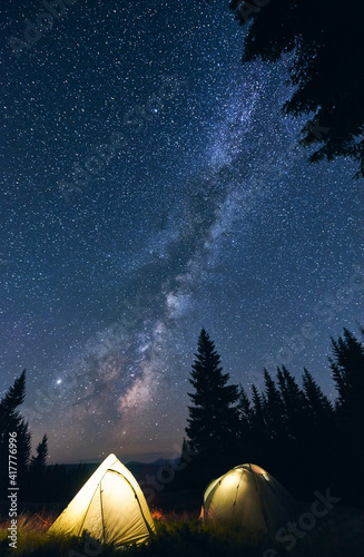 Vertical shot of tourist camping in forest, warm summer evening. Two illuminated tents under beautiful night sky full of stars and shiny milky way. Concept of camping in the mountains.