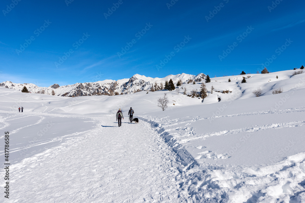 Hikers on a snowy footpath in winter landscape on the Lessinia Plateau (Altopiano della Lessinia), Regional Natural Park. On background the Monte Carega. Veneto and Trentino Alto Adige, Italy, Europe. Hikers on a snowy footpath in winter landscape on the Lessinia Plateau (Altopiano della Lessinia), Regional Natural Park. On background the Monte Carega. Veneto and Trentino Alto Adige, Italy, Europe.