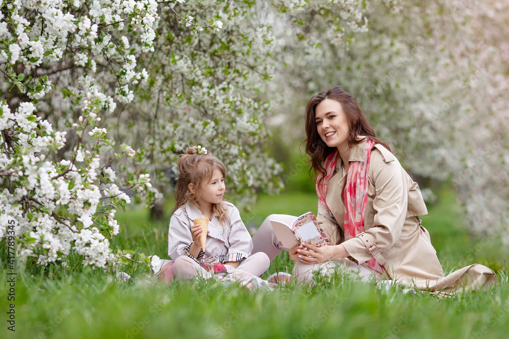 Mother and little daughter hugging each other and walking in blooming apple garden. Spring story. Happy family in beautiful spring day. Happy mother with daughter with book in spring garden Mother and little daughter hugging each other and walking in blooming apple garden. Spring story. Happy family in beautiful spring day. Happy mother with daughter with book in spring garden