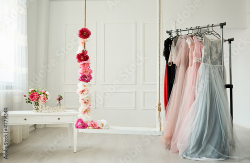 white wardrobe, large white strap, beautiful dresses in pastel colors hang on the hanger. Next to it, a rope entwined with beautiful, intense flowers.